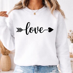 May include: White sweatshirt with the word "love" in black script, styled as an arrow. The arrow's head is a black heart. The sweatshirt is a casual, everyday garment.
