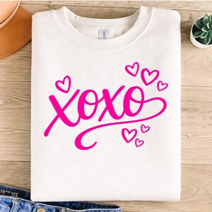 May include: A white sweatshirt with the word "xoxo" in bright pink script, surrounded by small pink hearts. The sweatshirt is folded neatly on a wooden surface, with a pair of jeans visible at the bottom.