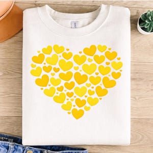 May include: White sweatshirt featuring a large heart design made of various sized yellow hearts. The sweatshirt is folded and placed on a wooden surface, with a pair of jeans visible at the bottom.
