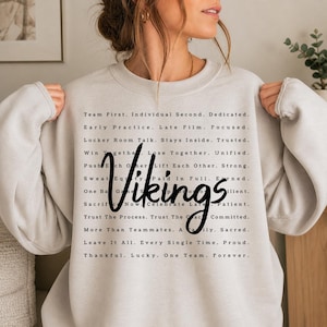 May include: A light grey crewneck sweatshirt featuring black text printed in a block layout. The word Vikings is written in a large, bold, cursive script across the centre, surrounded by rows of motivational phrases like Team First and Dedicated.