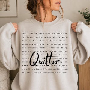 May include: A cream-colored sweatshirt with the word "Quitter" in a cursive font. The shirt features a list of quilting-related words and phrases in a black font, such as "Fabric Chosen," "Rotary Blade," and "Hand Quilted."