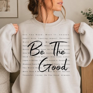 May include: Cream-colored sweatshirt with the words "Be The Good" in a black script font. The sweatshirt has a list of inspirational phrases in a smaller font. The overall design is simple and motivational.