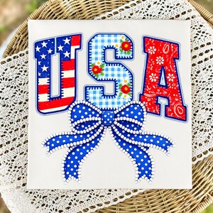 Op de afbeelding: Een patriottisch kunstwerk met de letters "USA" in rood, wit en blauw. De "U" heeft een sterrenpatroon, de "S" is ruitjes met bloemaccenten en de "A" is paisley. Onderaan een grote blauwe strik met witte stippen.