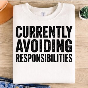 May include: White sweatshirt with the text "CURRENTLY AVOIDING RESPONSIBILITIES" in bold black letters. The sweatshirt is folded on a light wood surface, with a brown leather item and a small plant visible.