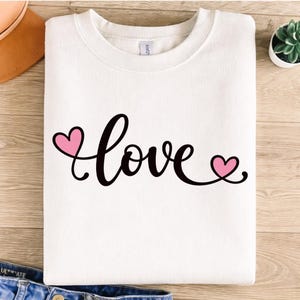 May include: White sweatshirt with the word "love" in black script, featuring two pink hearts. The sweatshirt is folded and placed on a wooden surface, with a pair of jeans visible at the bottom.