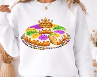 May include: White sweatshirt featuring a colorful illustration of a King Cake, a traditional pastry with a golden crown on top. The cake is decorated with purple, green, and gold sprinkles and a beaded necklace.