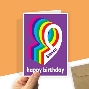 Op de afbeelding: Een rechthoekige wenskaart met een paarse achtergrond en een groot, gestileerd cijfer negen in regenboogkleuren. De naam Sienna is in het paars gedrukt op het witte gedeelte van het cijfer. De tekst happy birthday staat in het wit onderaan.