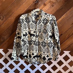 May include: Cream-colored button-down shirt with a black geometric and nature-themed pattern. The design includes snowflakes, trees, and animal silhouettes. The shirt has two chest pockets and long sleeves.