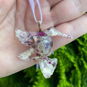 May include: A clear resin hummingbird pendant with dried flowers and a gold chain. The pendant is hanging from a light purple ribbon.