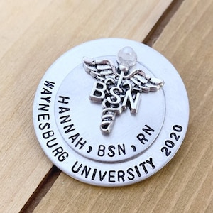 May include: Silver round charm with a caduceus symbol, the text "BSN, RN" and "HANNAH, BSN, RN 2020" and "WAYNESBURG UNIVERSITY".