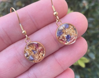 May include: Gold earrings with a round, clear resin pendant filled with dried flowers and gold flakes. The flowers are purple, pink, and yellow.
