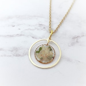 May include: A gold chain necklace with a delicate circle pendant. The pendant encloses a round resin charm with pressed white flowers and green leaves.