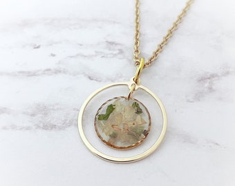 Op de afbeelding: Een gouden ketting met een delicate cirkelvormige hanger. De hanger omsluit een ronde harsbedel met geperste witte bloemen en groene bladeren.