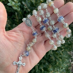 Puede incluir: Un rosario de plata con cuentas de cristal transparentes, amarillas y azules. La pieza central presenta un colgante de plata con una representaci&oacute;n de la Virgen Mar&iacute;a.