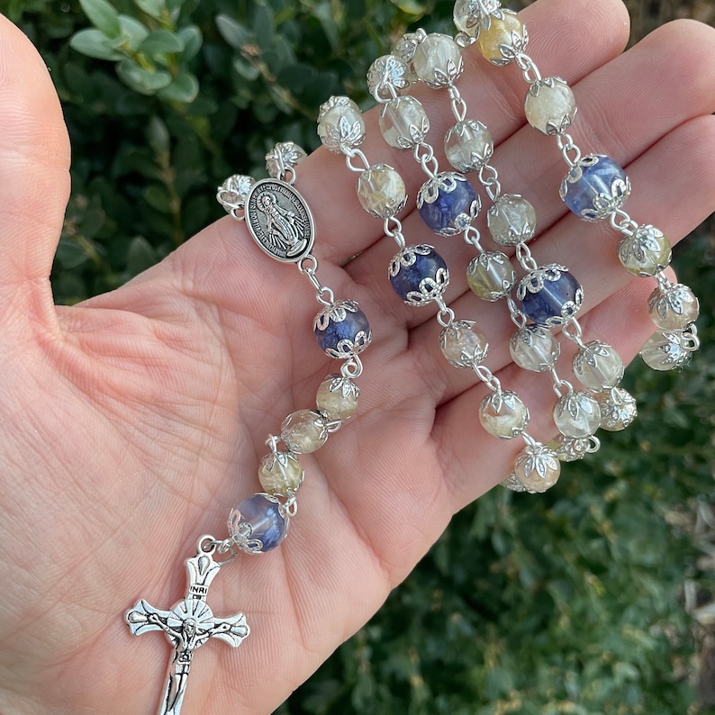 Puede incluir: Un rosario de plata con cuentas de cristal transparentes, amarillas y azules. La pieza central presenta un colgante de plata con una representaci&oacute;n de la Virgen Mar&iacute;a.