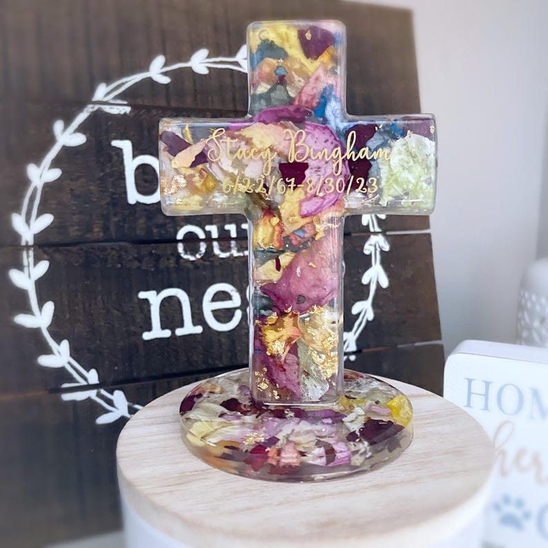 May include: A clear resin cross filled with dried flowers and gold flakes, set on a wooden base. The cross features the name "Stacy Bingham" and dates "6/22/67-8/30/23" in gold lettering. The cross is a memorial keepsake.