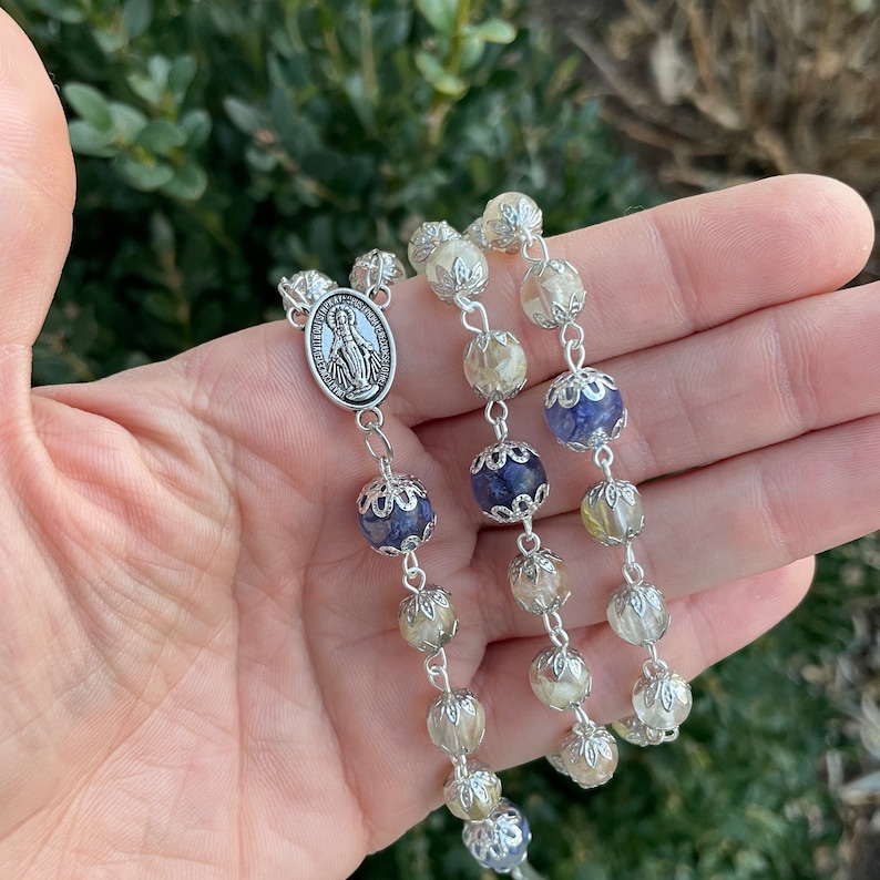 Puede incluir: Un rosario de plata con cuentas de cristal azul y transparente. La pieza central presenta un dije de plata de la Virgen Mar&iacute;a.