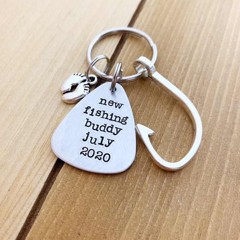 K&ouml;nnte beinhalten: Silberner Schl&uuml;sselanh&auml;nger mit einem Fischhaken-Anh&auml;nger und einem kleinen Anh&auml;nger mit einem Fu&szlig;abdruck-Design. Das Schild tr&auml;gt die Aufschrift "new fishing buddy july 2020".