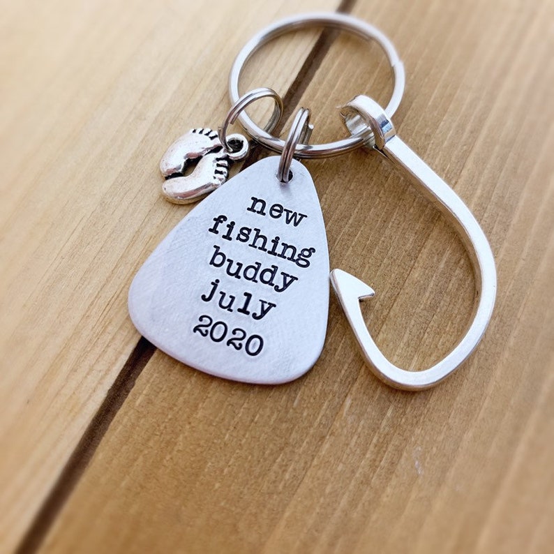 K&ouml;nnte beinhalten: Silberner Schl&uuml;sselanh&auml;nger mit einem Angelhaken-Anh&auml;nger und einem kleinen Anh&auml;nger mit Babyf&uuml;&szlig;en. Der Schl&uuml;sselanh&auml;nger hat ein silbernes Schild mit dem Text "new fishing buddy july 2020".