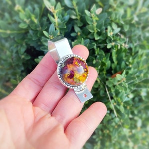 May include: A silver key chain with a round, clear resin charm containing pressed dried flowers in shades of red, yellow, and orange.