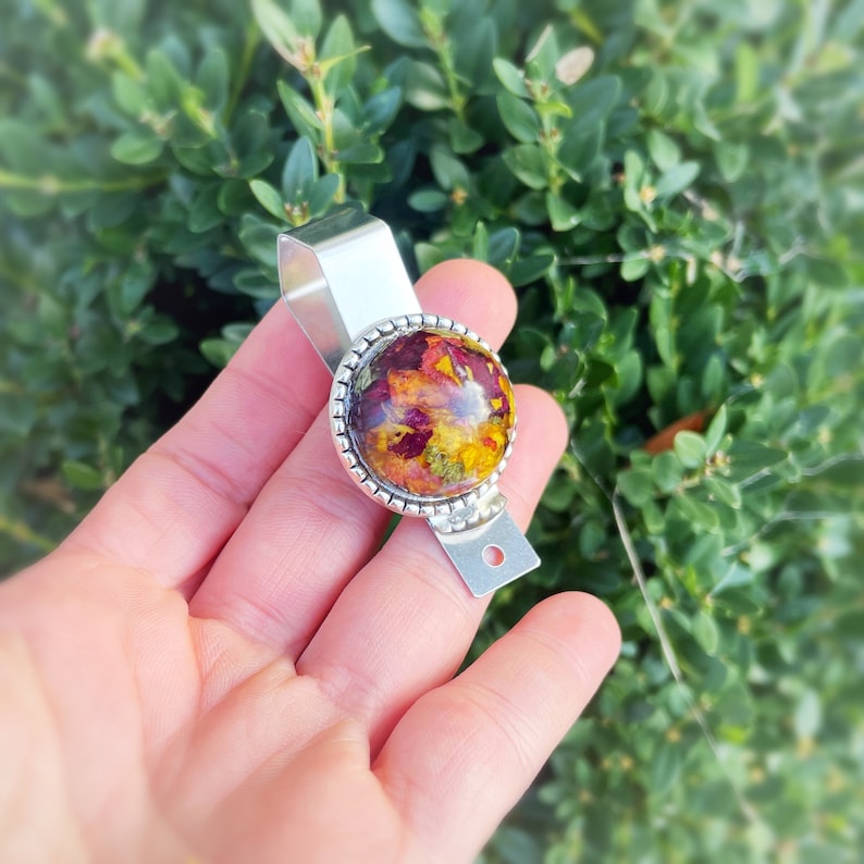 Puede incluir: Un llavero de plata con un charm redondo de resina transparente que contiene flores secas prensadas en tonos rojos, amarillos y naranjas.
