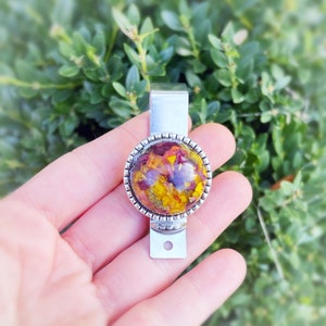 Puede incluir: Un llavero de metal plateado con un dije redondo de resina transparente que contiene flores prensadas en tonos amarillos, naranjas y rojos.