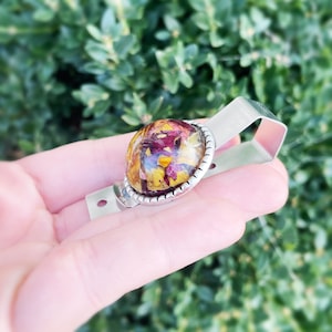 Puede incluir: Un porta credenciales de metal plateado con un bot&oacute;n redondo de resina transparente que presenta flores secas prensadas en tonos rojos, amarillos y naranjas.