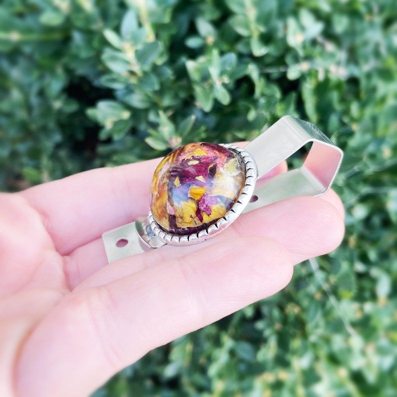 Puede incluir: Un porta credenciales de metal plateado con un bot&oacute;n redondo de resina transparente que presenta flores secas prensadas en tonos rojos, amarillos y naranjas.