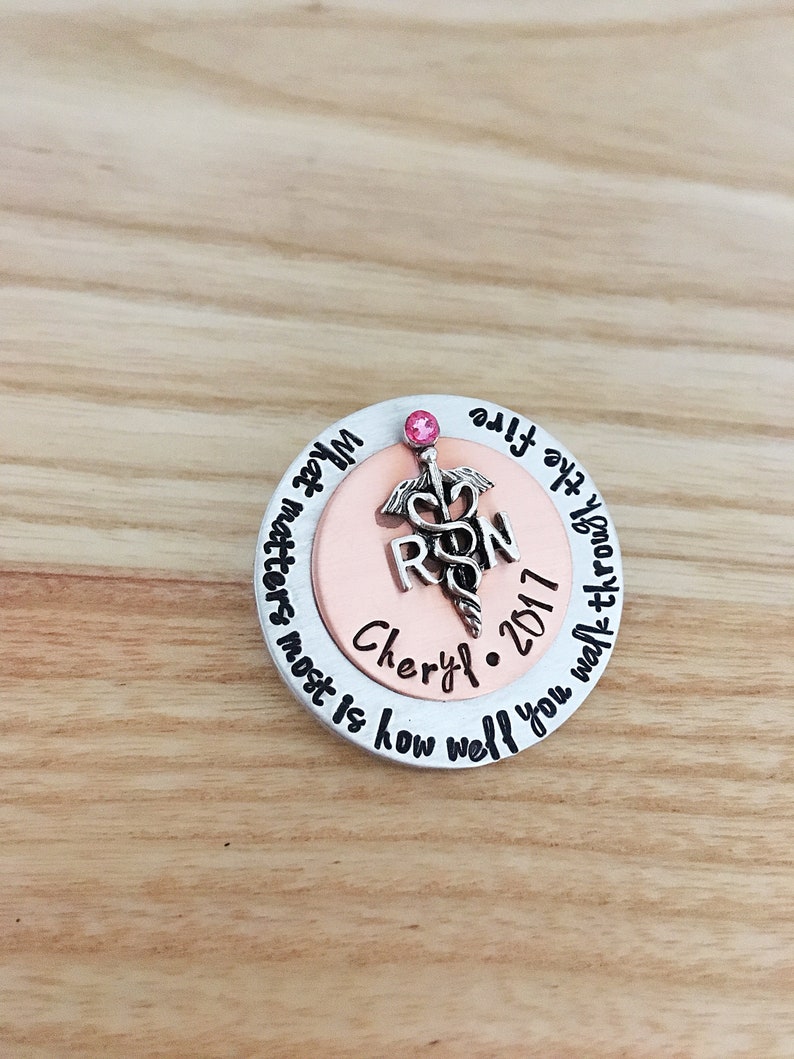May include: A silver and copper round pin with a pink rhinestone. The pin features a medical caduceus symbol with the letters "RN" and the text "Cheryl 2017". The text around the edge of the pin reads "How well you walk through life matters most is how well you walk through life".