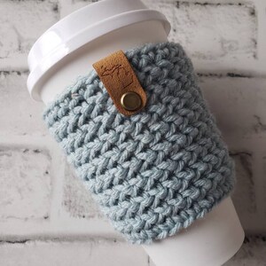 May include: A light blue crocheted coffee cup cozy with a brown leather strap and a brass button. The cozy is on a white to-go coffee cup.