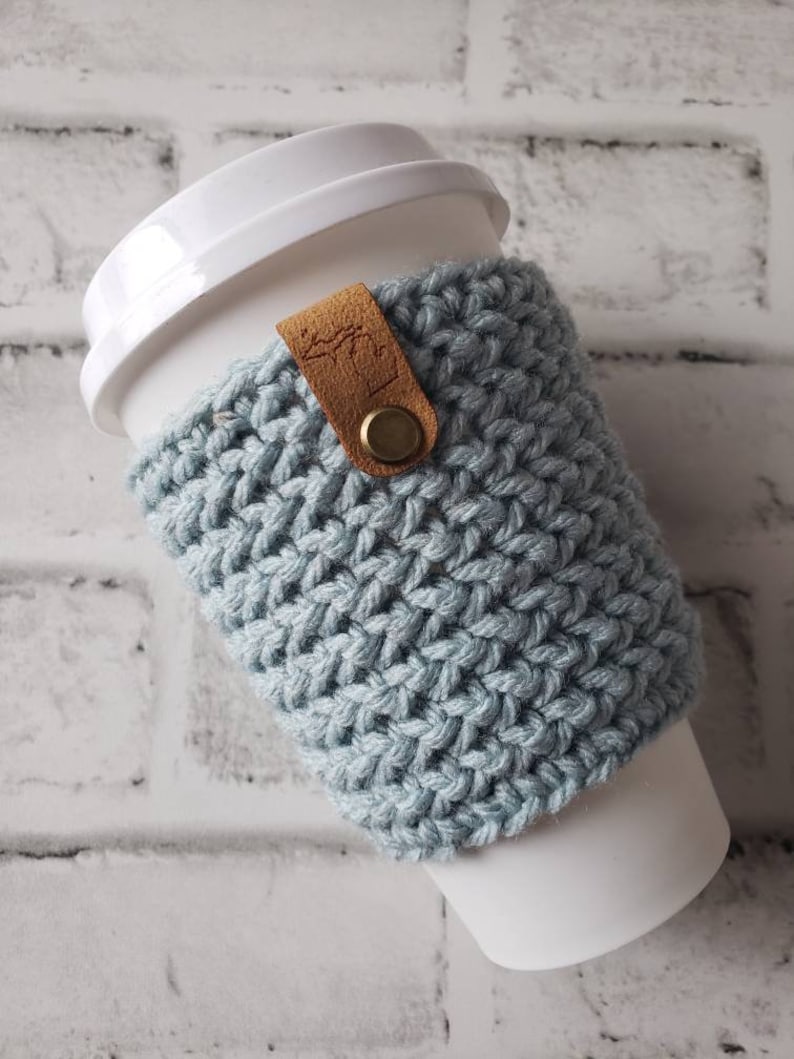 May include: A light blue crocheted coffee cup cozy with a brown leather strap and a brass button. The cozy is on a white to-go coffee cup.