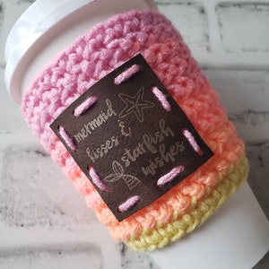 May include: A pink, orange, and yellow crocheted coffee cup cozy with a brown leather patch that says "mermaid kisses & starfish wishes".