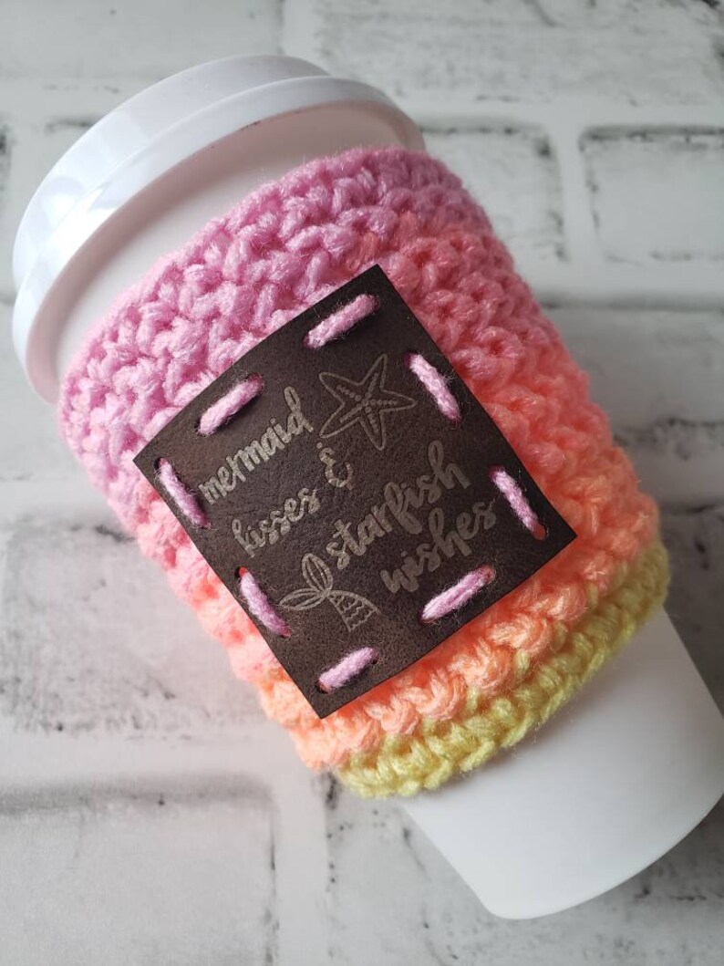May include: A pink, orange, and yellow crocheted coffee cup cozy with a brown leather patch that says "mermaid kisses & starfish wishes".