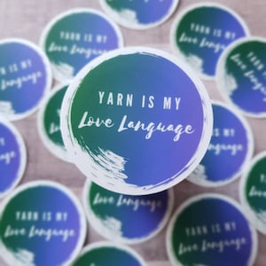 May include: A collection of round stickers with the text "Yarn is my Love Language." The stickers have a gradient background of blue and green with white text. The stickers are white with a brushstroke design.