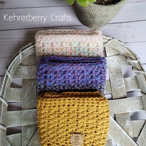 May include: Three crocheted wrist warmers stacked on a white wicker basket. The bottom warmer is mustard yellow, the middle warmer is purple and blue, and the top warmer is pink and white. A small brown tag with the text "Kehrberry Crafts" is attached to the bottom warmer.