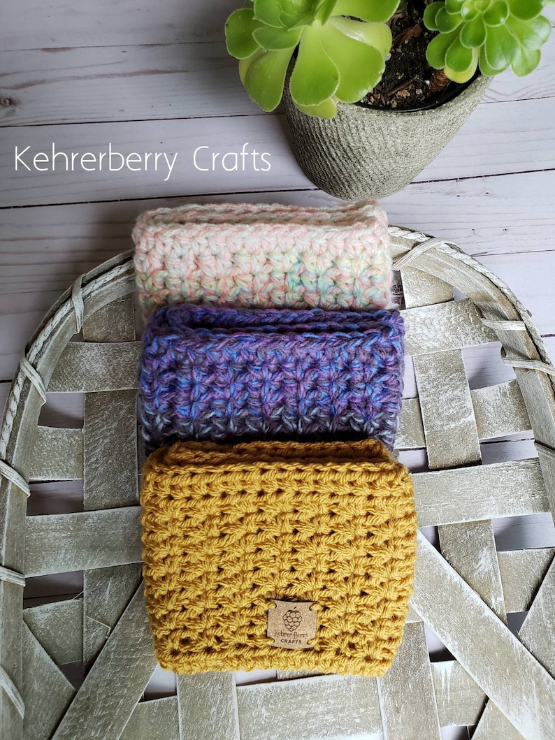 May include: Three crocheted wrist warmers stacked on a white wicker basket. The bottom warmer is mustard yellow, the middle warmer is purple and blue, and the top warmer is pink and white. A small brown tag with the text "Kehrberry Crafts" is attached to the bottom warmer.