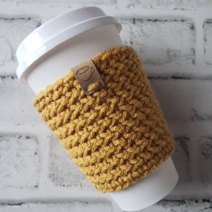May include: A yellow crocheted coffee cup cozy with a brown leather tag. The cozy is on a white to-go cup.