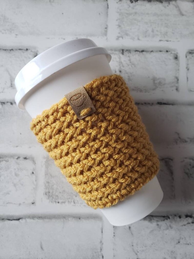 May include: A yellow crocheted coffee cup cozy with a brown leather tag. The cozy is on a white to-go cup.