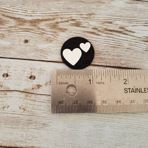 Puede incluir: Un c&iacute;rculo de fieltro negro con dos corazones blancos recortados. El c&iacute;rculo tiene un di&aacute;metro de aproximadamente 2,5 cm.
