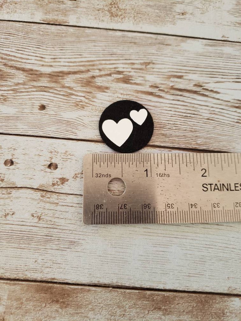 Puede incluir: Un c&iacute;rculo de fieltro negro con dos corazones blancos recortados. El c&iacute;rculo tiene un di&aacute;metro de aproximadamente 2,5 cm.