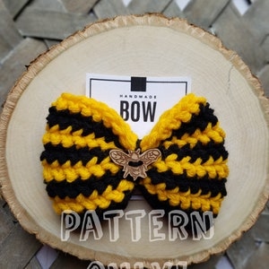 Puede incluir: Un lazo de crochet a rayas amarillas y negras con un dije de abeja de madera. El lazo está sobre una superficie de madera con una etiqueta blanca que dice "Handmade Bow" y el texto "PATTERN ONLY!" está superpuesto en la imagen.