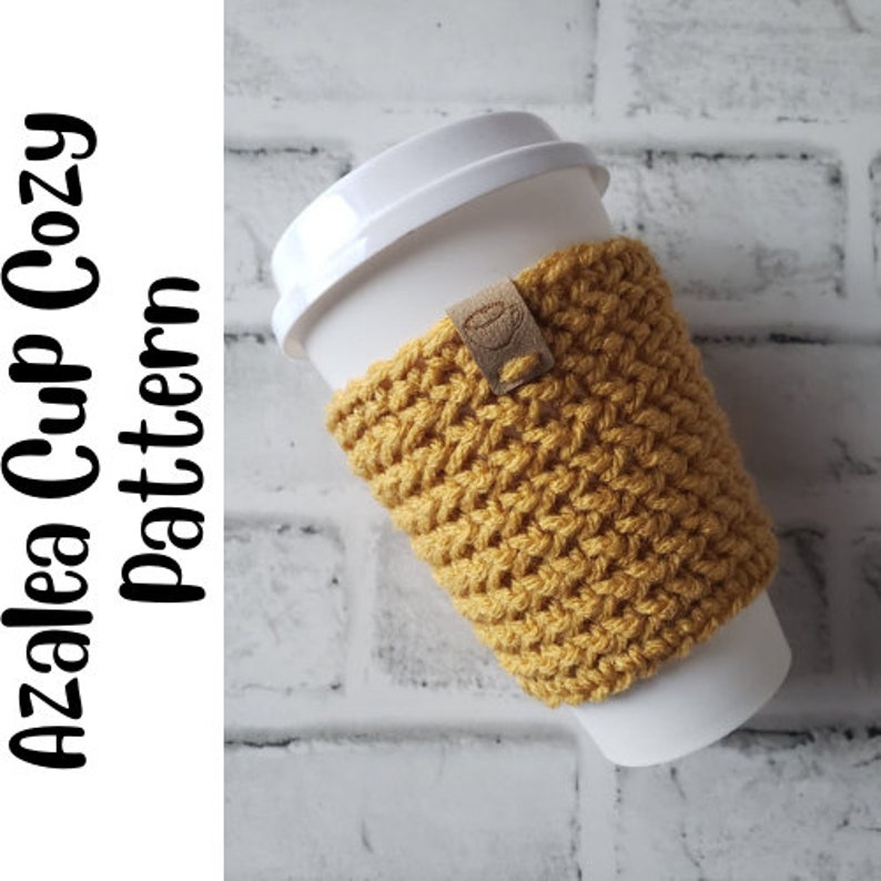 May include: A crocheted cup cozy in a mustard yellow color with a brown leather tag. The cozy is on a white to-go cup.