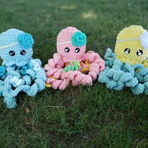May include: Three crocheted octopus plushies in different colors: light blue, pink, and yellow. Each octopus has a crocheted flower on its head and large, black eyes.