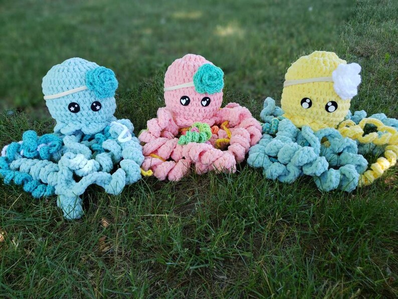 May include: Three crocheted octopus plushies in different colors: light blue, pink, and yellow. Each octopus has a crocheted flower on its head and large, black eyes.