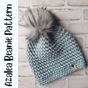 May include: A light blue crocheted beanie with a white and black faux fur pom pom. The beanie has a small brown leather tag with the text "The Hook Nook" on it.