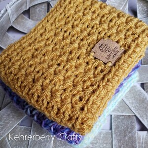May include: A stack of crocheted blankets in shades of yellow, purple, and blue. The top blanket is a solid yellow and features a brown leather tag with the text "Kehrerberry Crafts" and a blackberry design.