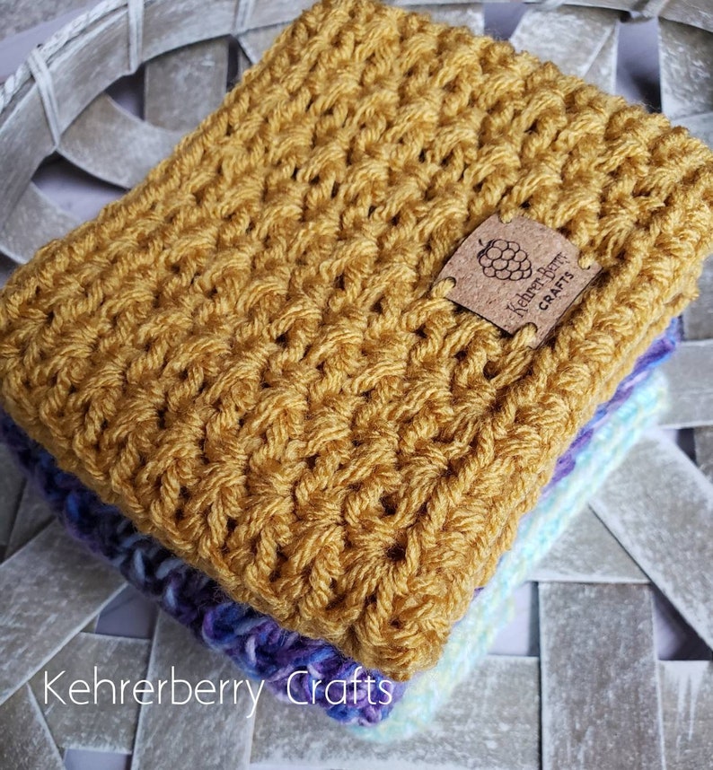May include: A stack of crocheted blankets in shades of yellow, purple, and blue. The top blanket is a solid yellow and features a brown leather tag with the text "Kehrerberry Crafts" and a blackberry design.
