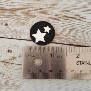 Puede incluir: Un c&iacute;rculo negro con dos estrellas blancas sobre &eacute;l. El c&iacute;rculo tiene un di&aacute;metro de aproximadamente 2,5 cm. El c&iacute;rculo est&aacute; sobre una superficie de madera.