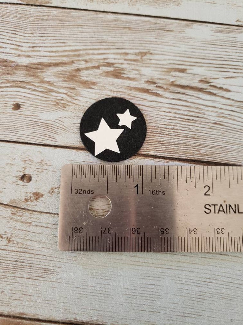 Puede incluir: Un c&iacute;rculo negro con dos estrellas blancas sobre &eacute;l. El c&iacute;rculo tiene un di&aacute;metro de aproximadamente 2,5 cm. El c&iacute;rculo est&aacute; sobre una superficie de madera.