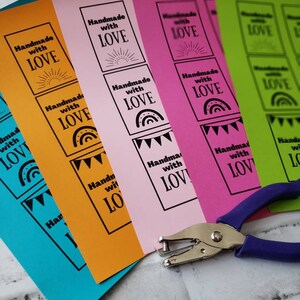 Pode incluir: Cinco folhas de papel com fundos de cores diferentes, cada uma com o texto "Handmade with Love" em letras pretas. O texto está em um quadrado com um design diferente em cada quadrado. O papel está empilhado um sobre o outro e um furador está em primeiro plano.